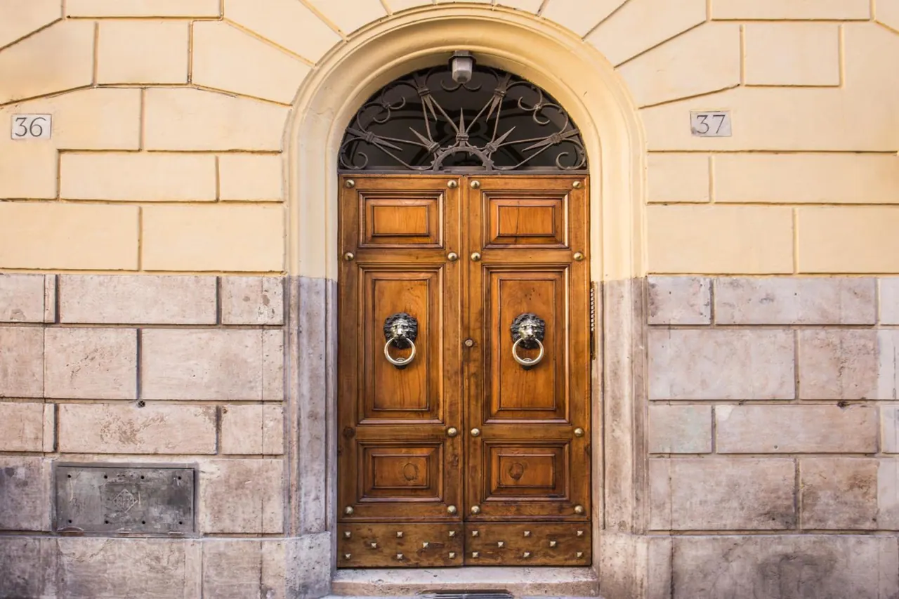 A wooden double door with lion-head knockers and an ornate ironwork fanlight sits in an arched doorway in a cream and stone facade, with house numbers 36 and 37 on the wall.