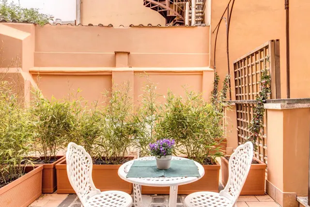 A small terrace with a white cast-metal table and two chairs, bamboo planters along an ochre wall, and a potted purple plant on the table.