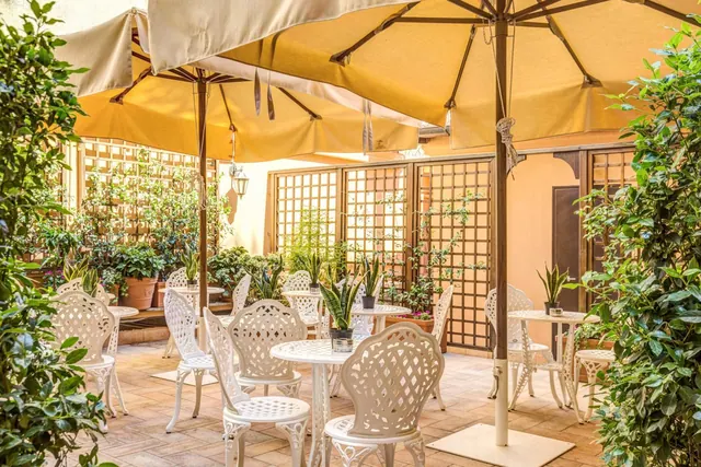 An outdoor courtyard dining area with white cast-iron chairs and tables under yellow parasols, surrounded by potted plants and lattice screens.