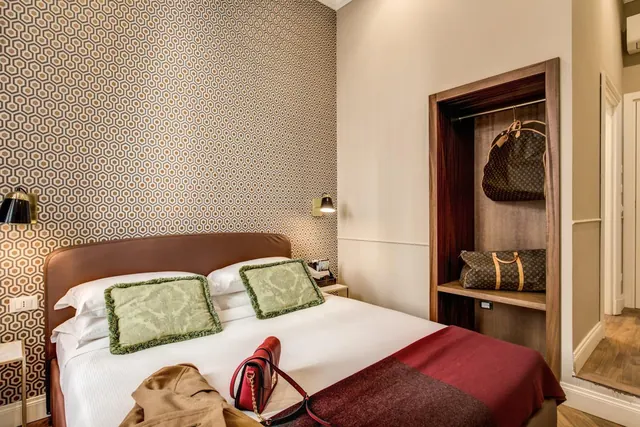 A double bed with green cushions and a red bag on it sits beside an open wardrobe holding luggage, in a room with a patterned wallpaper wall.