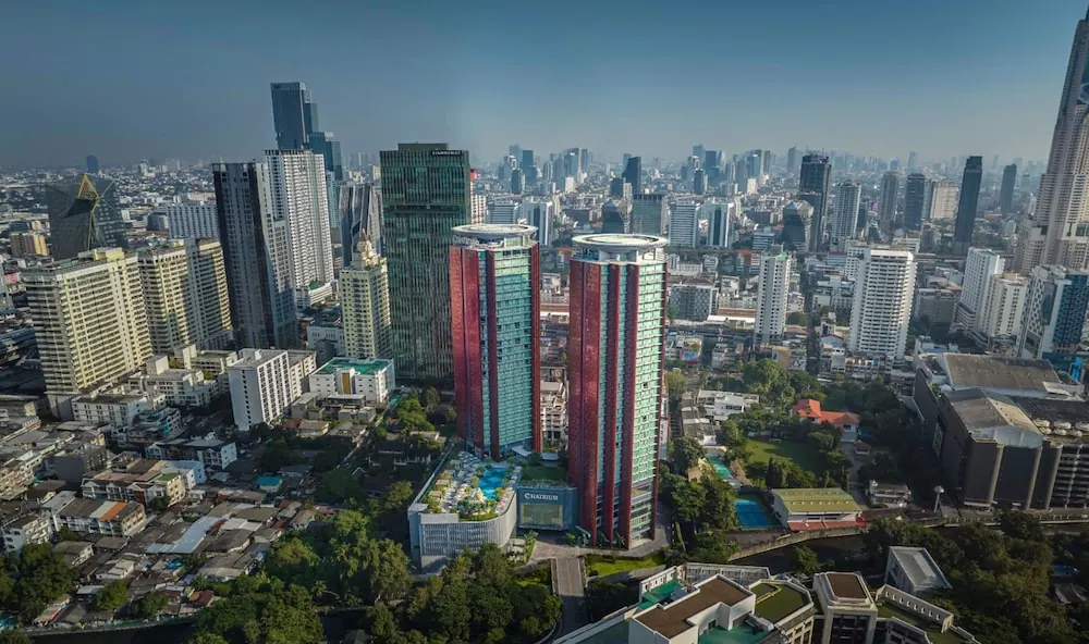 Aerial view of the Chatrium hotel's twin red and green glass towers and a low podium with a rooftop pool, surrounded by Bangkok's high-rise skyline.