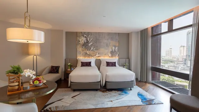A hotel bedroom with two single beds, a seating area, a round table with fruit and orchids, and a large window overlooking city buildings.