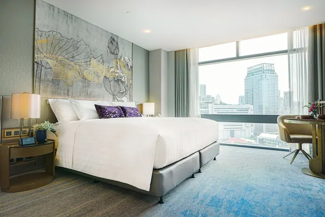 A bedroom with a king bed, a large framed artwork on the wall, a window overlooking high-rise buildings, and a work desk with a chair on the right.
