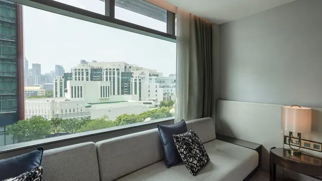 A living area with a grey sofa and cushions beside a large window overlooking city buildings and trees, with a table lamp on the right.