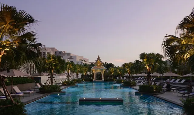 Evening view of a large outdoor swimming pool lined with sun loungers and palm trees, with a golden Thai-style pavilion at the far end.