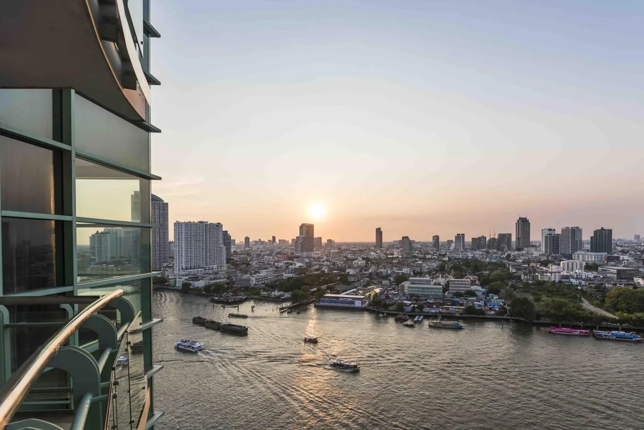 A sunset view over the Chao Phraya river and city skyline from a high-floor balcony, with boats on the water.
