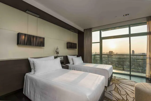 A bedroom with two made beds facing floor-to-ceiling windows that look out over a city skyline at sunset.