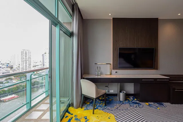 A hotel room with a wall-mounted television, a desk with a chair, and sliding glass doors opening onto a balcony overlooking a river and city buildings.