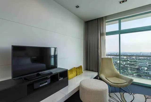 A living area with a flat-screen television on a black console, a white built-in bench with yellow cushions, an armchair and an ottoman, and floor-to-ceiling windows overlooking a city.