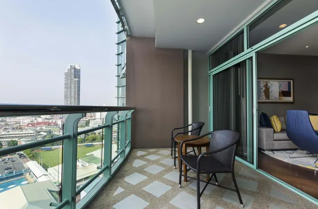 A furnished balcony with two dark wicker chairs and a small round table overlooks a city with a tall tower and a sports field, with a sofa and blue chair visible through the glass doors.