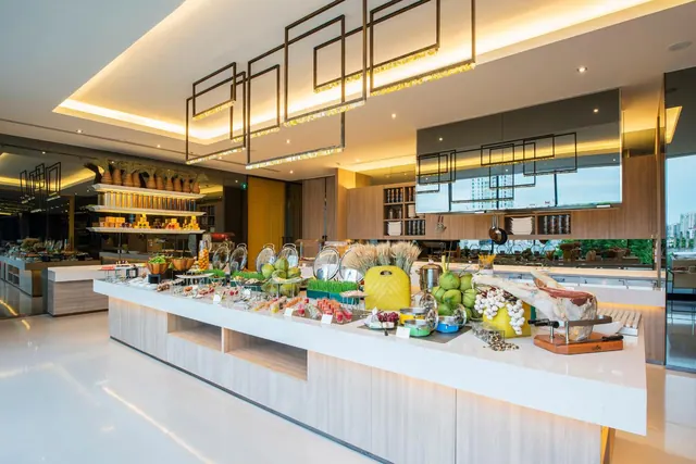 A buffet counter with food displays, a ham carving station and green coconuts sits under geometric ceiling lights in a restaurant, with high-rise buildings visible through the window on the right.