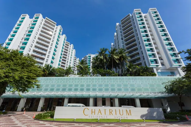 Exterior of the Chatrium Residence Sathon, showing tall white towers, a glass canopy over the entrance, and a sign reading Chatrium Residence Sathon under a clear blue sky.