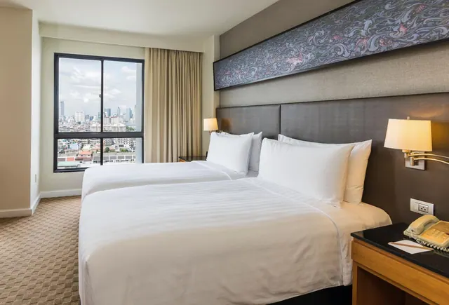 A bedroom with two made beds under a long framed wall artwork, a bedside lamp and telephone, and a window looking out over a city skyline.