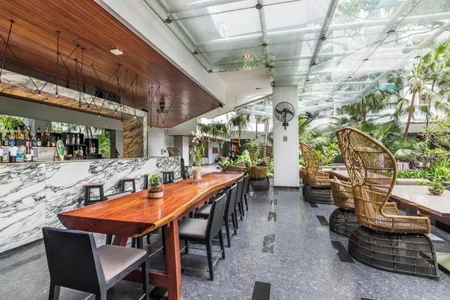 A covered lounge area with a long wooden bar table, dark chairs, wicker lounge chairs and a marble bar counter beneath a glass roof with palm trees.