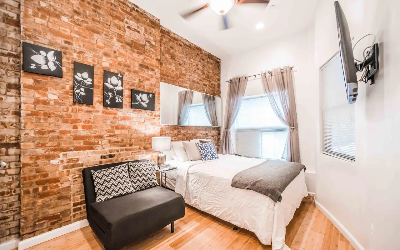 A bedroom with an exposed brick wall, a double bed with white bedding, a dark armchair, a wall-mounted television and a ceiling fan.