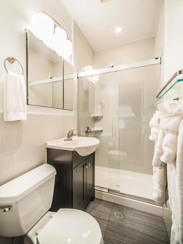 A bathroom with a single sink vanity, a toilet, and a walk-in shower enclosed by glass with a sliding door, with white towels hanging nearby.