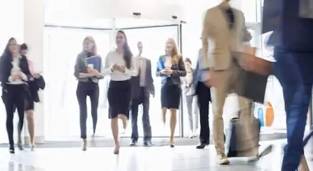Several businesspeople walk through a bright lobby past a revolving door, with one man in a tan suit carrying a bag in the foreground.