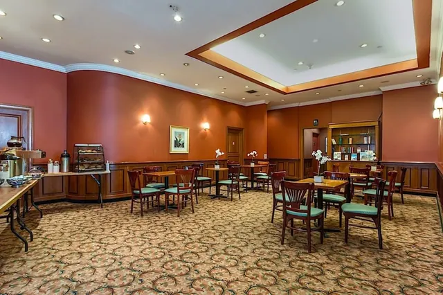 A dining room with wooden chairs and small tables, a buffet counter with coffee urns and a pastry display case on the left, and orchids on some of the tables.