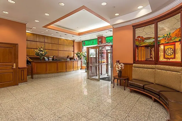 A hotel lobby with a wooden reception desk and two staff behind it, a curved leather bench beneath stained glass windows, and glass entrance doors.