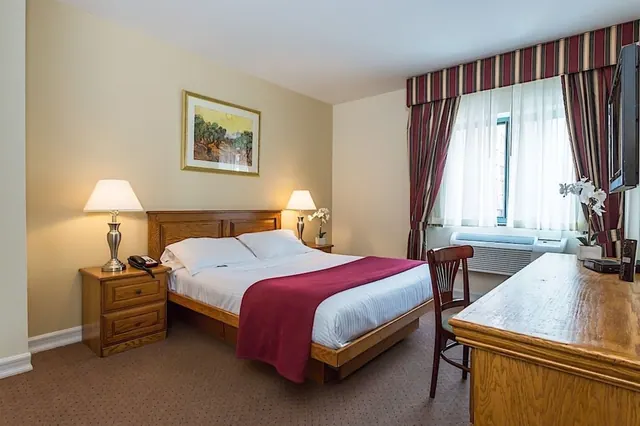 A double bed with a red throw and two bedside lamps sits beside a wooden desk and wall-mounted television in a hotel bedroom.