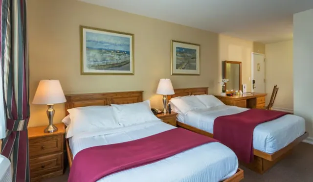 A hotel bedroom with two double beds with wooden headboards, bedside lamps, framed artwork on the wall and a wooden desk with a mirror on the right.