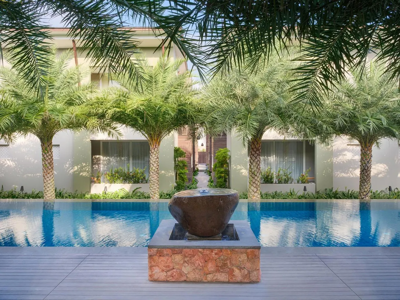 A long swimming pool with a dark stone water bowl fountain on a stone base, with palm trees and a white building with windows behind it.