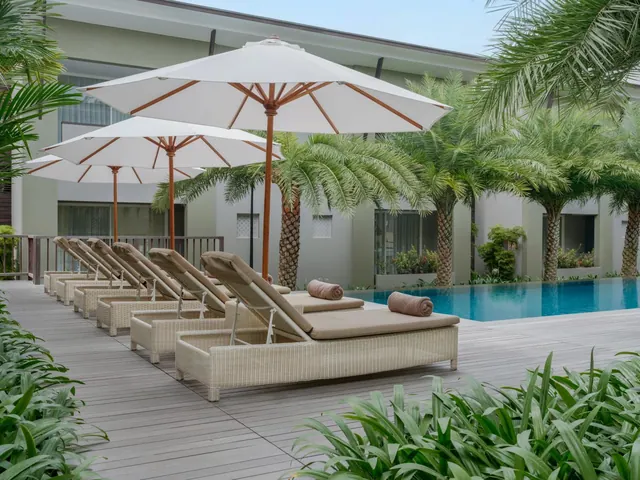 A row of wicker sun loungers with rolled towels and white parasols stands on a timber deck beside a swimming pool, with palm trees behind.