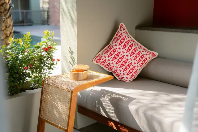 A cushioned daybed with a red patterned pillow sits beside a cane side table holding a wooden dish, with flowering plants and a pool visible through the open doorway.