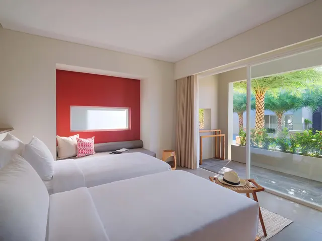 A bedroom with two white beds, a red accent wall with a built-in bench, and a sliding glass door looking onto a terrace with palm trees and garden planting.