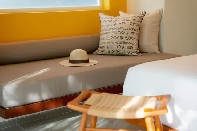 A cushioned built-in bench under a yellow-walled window holds a straw hat and patterned pillows, with a woven wooden stool in the foreground and a white bed at right.