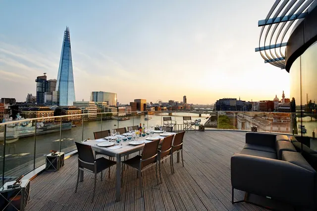 A rooftop terrace with a laid dining table and outdoor sofa overlooks the River Thames and The Shard at sunset.