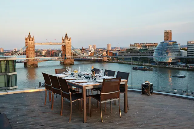 A set dining table and chairs on a rooftop terrace overlooking the River Thames, with Tower Bridge and City Hall visible.