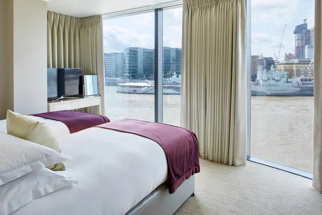 A bedroom with two single beds with burgundy throws and a television, with floor-to-ceiling windows overlooking the River Thames, a moored warship and city buildings.