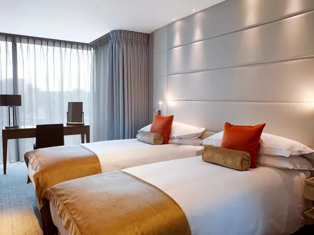 A bedroom with two single beds with white bedding, tan runners and orange cushions, beside a desk and chair near a window with sheer curtains.
