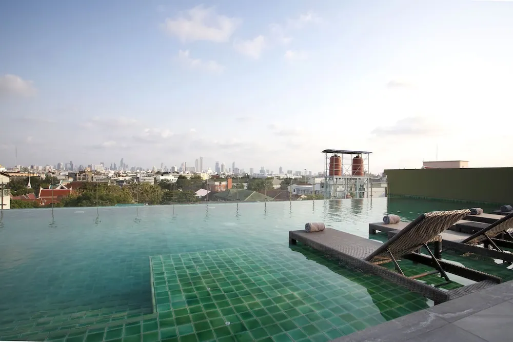 A rooftop infinity pool with green tiles overlooks a distant city skyline, with sun loungers and rolled towels on the right.