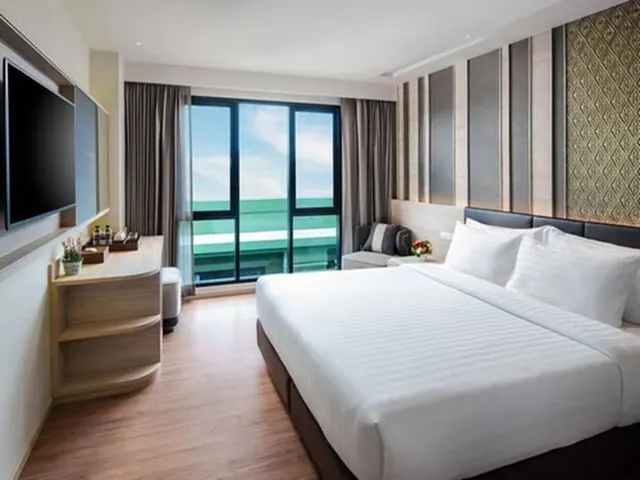 A bedroom with a made-up king bed, a wall-mounted television, a wooden cabinet with shelves, and a small seating area beside a window with curtains.