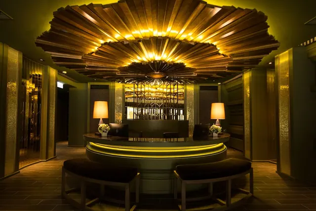 A hotel lobby with a curved reception desk, a fan-shaped wooden ceiling lit in gold, two table lamps with white flower arrangements, and two low stools in the foreground.