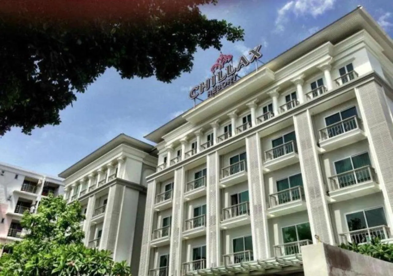 A low-angle view of a white multi-storey hotel building with rows of balconies and a rooftop sign reading Chillax Heights, with trees and blue sky visible.