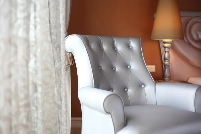 A white tufted leather armchair sits beside a sheer curtain and a lit table lamp against an orange wall.