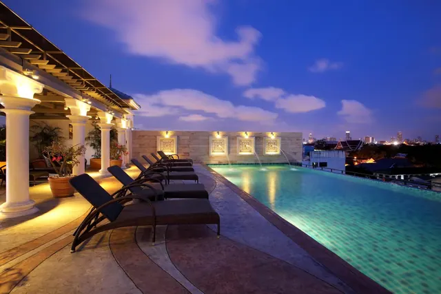 A rooftop swimming pool at dusk with lounge chairs along its edge, white columns on the left and a city skyline in the distance.