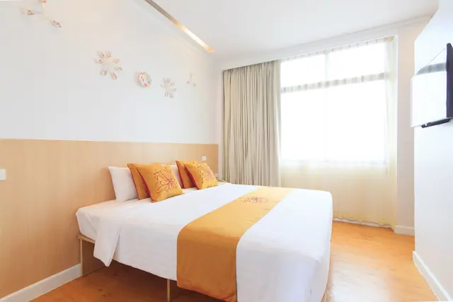 A bedroom with a double bed dressed in white linen and orange cushions, a wall-mounted television, and a window with sheer curtains.