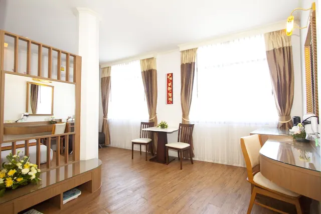 A bright hotel room with a small wooden dining table and two chairs in front of tall curtained windows, a glass-topped vanity desk on the right, and a flower arrangement on a wooden divider in the foreground.