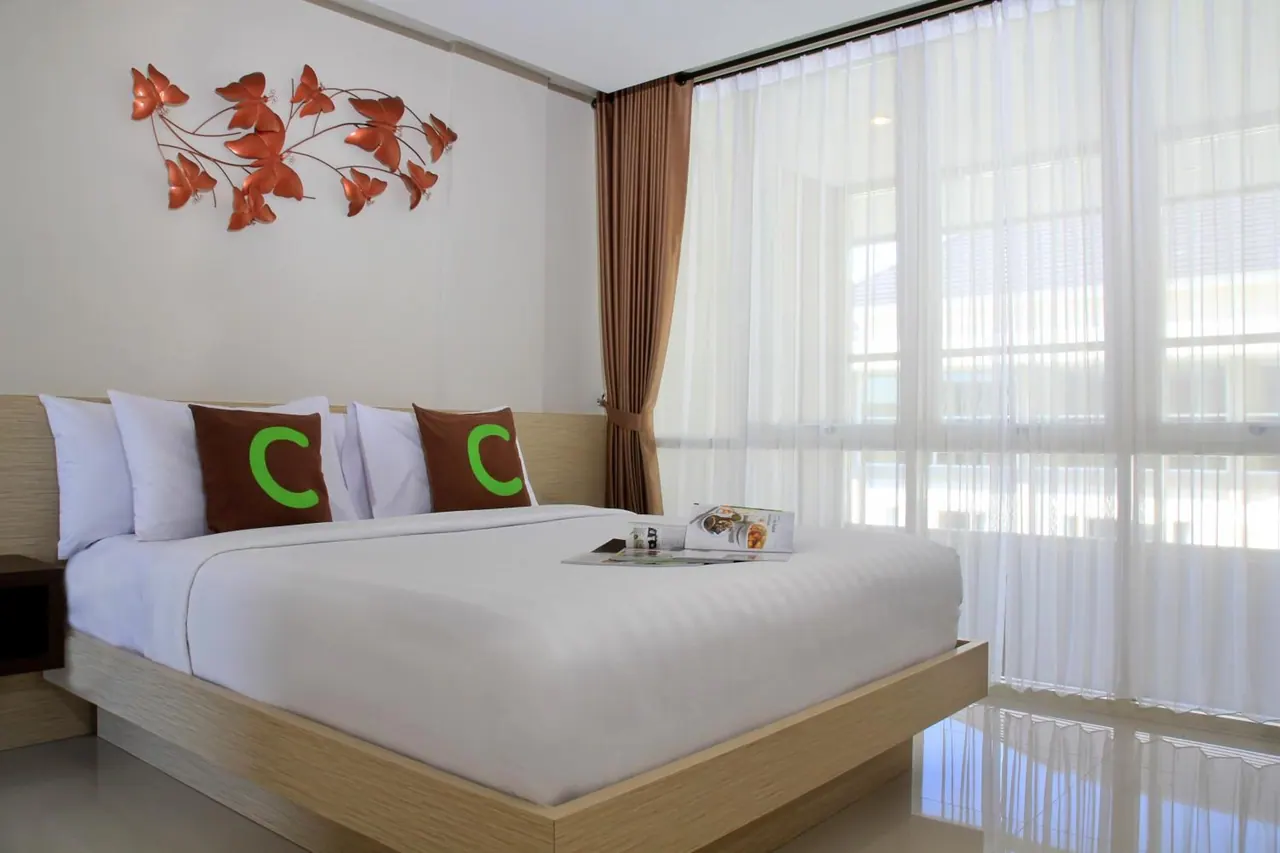 A king bed with white linens, brown pillows and a magazine sits beside sheer-curtained floor-to-ceiling windows, with a metal butterfly wall decoration above the headboard.