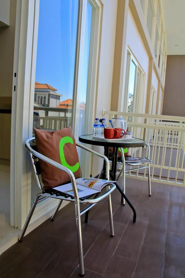 A narrow balcony with two aluminium chairs and a round table holding a tray with red mugs, water bottles and a kettle, beside a glass door.