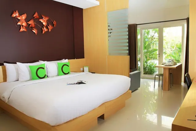 A bedroom with a king bed, white bedding and green cushions, a copper butterfly wall decoration, and a small table by glass doors opening onto a garden.