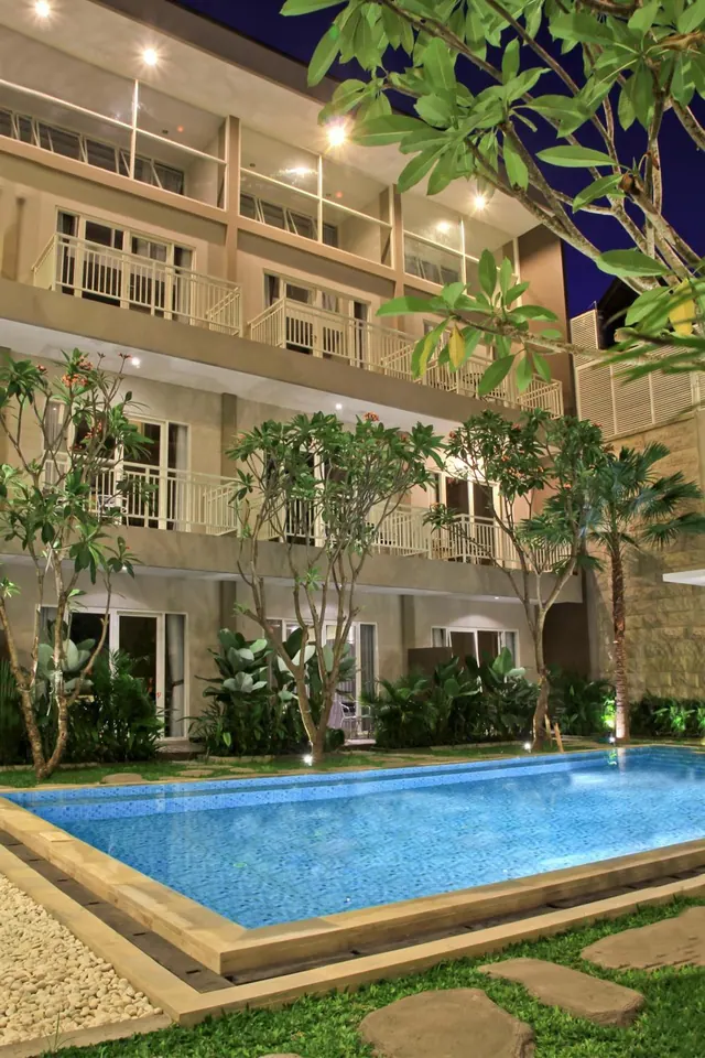A rectangular swimming pool sits in front of a multi-storey hotel building with balconies, lit at night, with frangipani trees and a landscaped garden beside it.