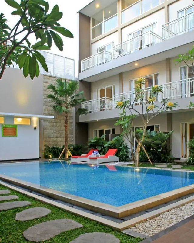 A rectangular swimming pool in a courtyard with white and red sun loungers, frangipani trees, and a multi-storey building with balconies behind it.