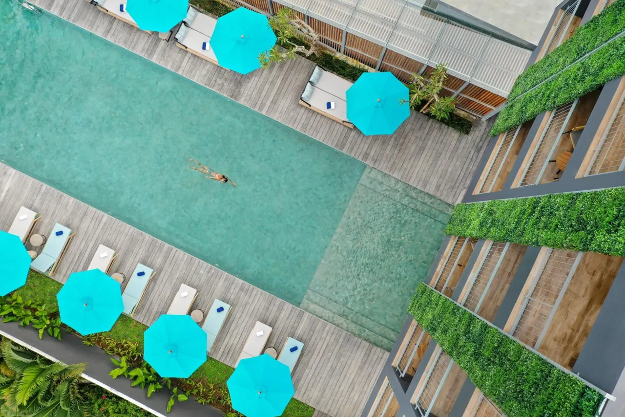 Aerial view of a swimming pool with turquoise parasols, sun loungers on timber decking, one swimmer, and green planted walls on the adjacent building.