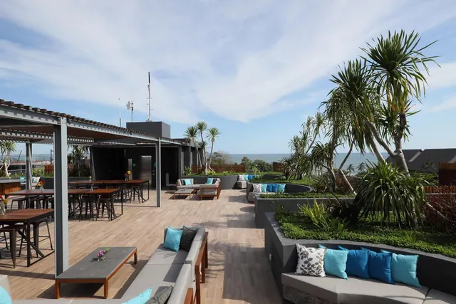 A rooftop terrace with lounge sofas, dining tables under a pergola, palm trees and the sea visible in the distance.