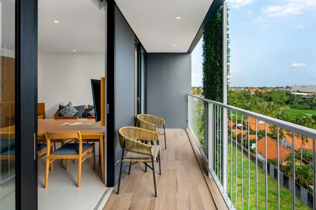 A private balcony with wooden flooring and two woven chairs, with the living room visible through sliding glass doors and a view over rooftops and green fields.